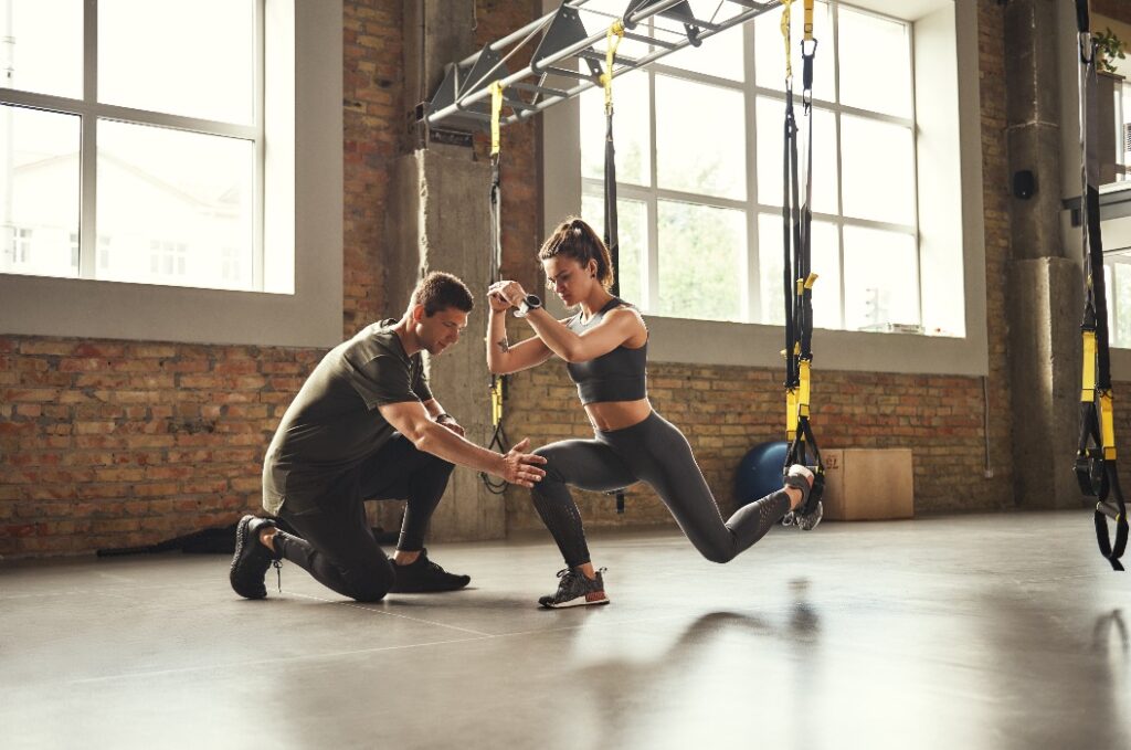 Kniebeugen üben. Ein selbstbewusster junger Personal Trainer zeigt einer schlanken, athletischen Frau beim Training im Fitnessstudio, wie man mit TRX-Fitnessbändern Kniebeugen ausführt.
