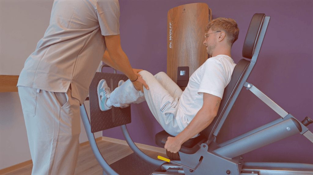 Therapeut unterstuetzt laechelnden jungen Mann bei Beinuebung an Beinpresse